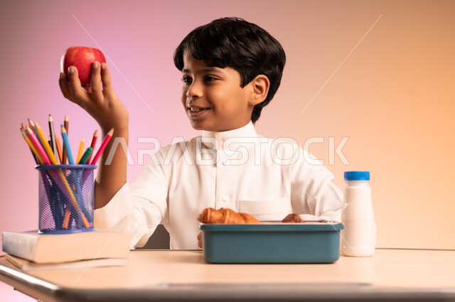 لانش بوكس فيه سناكات صحية مغذية، موسم العودة إلى المدارس في السعودية، إيماءات السعادة والسرور، القرطاسية المدرسية والأقلام والكتب العلمية، بورتريه لطالب عربي خليجي سعودي يرتدي الثوب التقليدي يجلس في مقعده يمسك بيده تفاحة من علبة الطعام أمامه،  خلفية ملونة