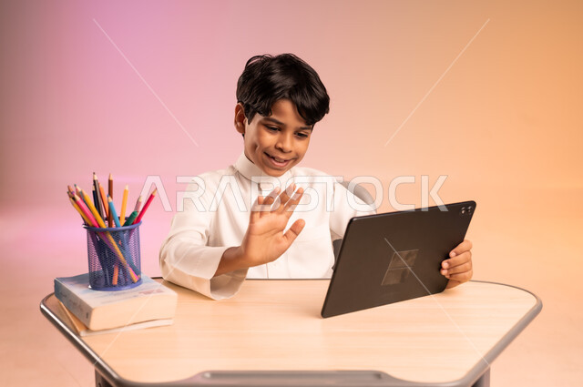 موسم العودة إلى المدارس، طرائق تعليمية جديدة متطورة، بورتريه لطالب عربي خليجي سعودي يرتدي الثوب التقليدي يجلس في مقعده يمسك بالجهاز اللوحي ويتابع الدروس عبر الانترنت بإيماءات الاندماج والتركيز، دمج التكنولوجيا بالتعليم في المدارس الحديثة، خلفية ملونة