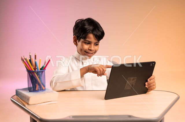 موسم العودة إلى المدارس، طرائق تعليمية جديدة متطورة، بورتريه لطالب عربي خليجي سعودي يرتدي الثوب التقليدي يجلس في مقعده يمسك بالجهاز اللوحي ويتابع الدروس عبر الانترنت بإيماءات الاندماج والتركيز، دمج التكنولوجيا بالتعليم في المدارس الحديثة، خلفية ملونة