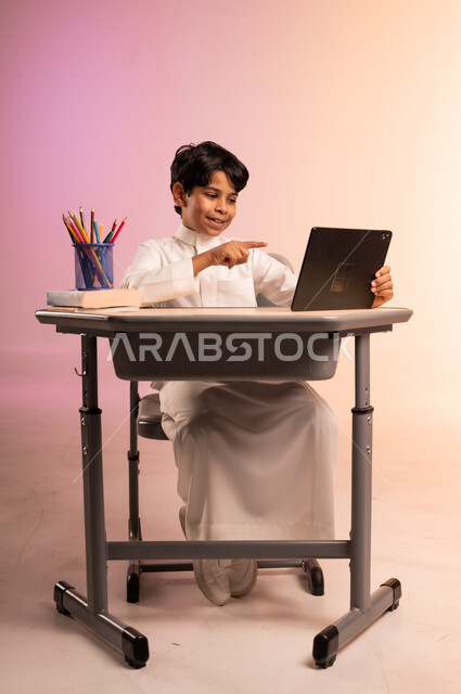 موسم العودة إلى المدارس، طرائق تعليمية جديدة متطورة، بورتريه لطالب عربي خليجي سعودي يرتدي الثوب التقليدي يجلس في مقعده يمسك بالجهاز اللوحي ويتابع الدروس عبر الانترنت بإيماءات الاندماج والتركيز، دمج التكنولوجيا بالتعليم في المدارس الحديثة، خلفية ملونة