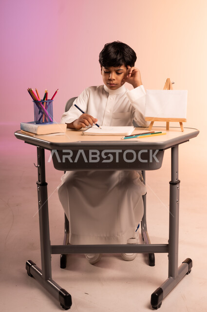  إيماءات الاندماج والتركيز في حل وكتابة الواجبات، مراجعة الدروس بجد واجتهاد للوصول إلى النجاح والتفوق، العودة إلى المدارس، بورتريه لطالب عربي خليجي سعودي يرتدي الثوب التقليدي يجلس في مقعده، قرطاسية مدسية ولوحة بيضاء فارغة على الطاولة المكتبية، خلفية ملونة