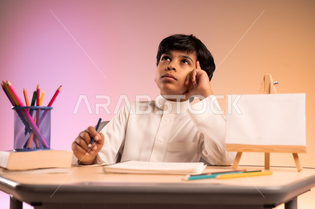  إيماءات الاندماج والتركيز في حل وكتابة الواجبات، مراجعة الدروس بجد واجتهاد للوصول إلى النجاح والتفوق، العودة إلى المدارس، بورتريه لطالب عربي خليجي سعودي يرتدي الثوب التقليدي يجلس في مقعده، قرطاسية مدسية ولوحة بيضاء فارغة على الطاولة المكتبية، خلفية ملونة