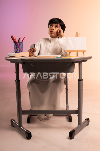 Gestures of integration and concentration in solving and writing assignments, reviewing lessons diligently and diligently to achieve success and excellence, back to school, portrait of a Saudi Arabian Gulf student wearing traditional dress sitting in his seat, stationery and an empty white board on the office table, colorful background