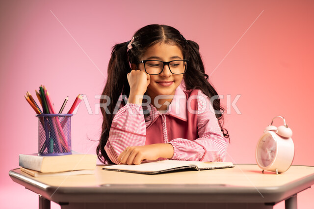 الجد والاجتهاد للوصول إلى النجاح والتفوق، موسم العودة إلى المدارس، بورتريه لطالبة عربية خليجية سعودية ترتدي الزي المدرسي وتضع نظارة طبية تجلس في مقعدها تقوم بالنظر إلى ساعة المنبه بسعادة، قرطاسية مدرسية على الطاولة المكتبية، خلفية ملونة