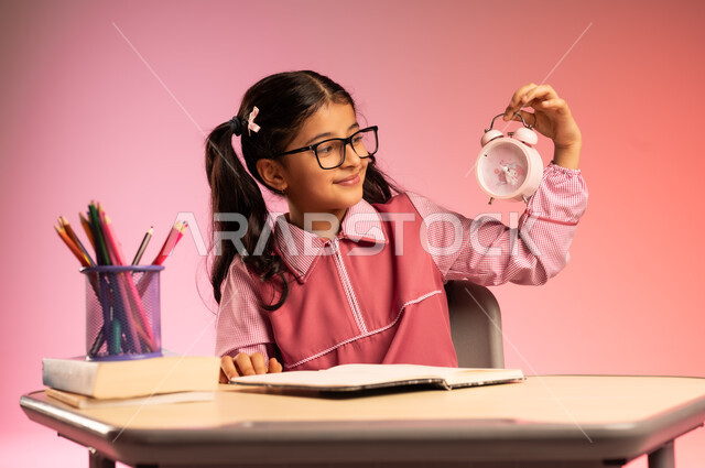 الجد والاجتهاد للوصول إلى النجاح والتفوق، موسم العودة إلى المدارس، بورتريه لطالبة عربية خليجية سعودية ترتدي الزي المدرسي وتضع نظارة طبية تجلس في مقعدها تقوم بالنظر إلى ساعة المنبه بسعادة، قرطاسية مدرسية على الطاولة المكتبية، خلفية ملونة