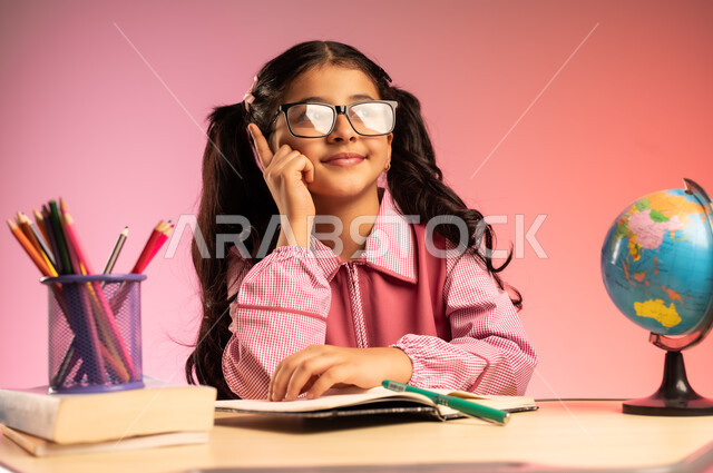 الاجتهاد للوصول إلى النجاح والتفوق، موسم العودة إلى المدارس، بورتريه لطالبة عربية خليجية سعودية ترتدي الزي المدرسي ونظارة طبية تجلس في مقعدها تقوم بالنظر إلى الأعلى بإيماءات الاندماج والسعادة، قرطاسية مدرسية ومجسم كرة أرضية على الطاولة المكتبية، خلفية ملونة
