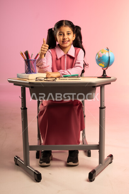 الاجتهاد للوصول إلى النجاح والتفوق، موسم العودة للمدارس، بورتريه لطالبة عربية خليجية سعودية ترتدي الزي المدرسي تجلس في مقعدها تنظر إلى الكاميرا بإيماءات الاندماج والسعادة، حل الواجبات والمراجعة، قرطاسية مدرسية ومجسم كرة أرضية على الطاولة المكتبية، خلفية ملونة