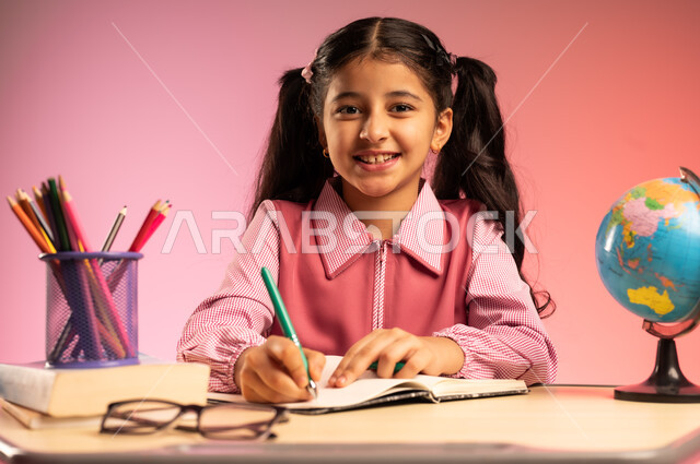 الاجتهاد للوصول إلى النجاح والتفوق، موسم العودة للمدارس، بورتريه لطالبة عربية خليجية سعودية ترتدي الزي المدرسي تجلس في مقعدها تنظر إلى الكاميرا بإيماءات الاندماج والسعادة، حل الواجبات والمراجعة، قرطاسية مدرسية ومجسم كرة أرضية على الطاولة المكتبية، خلفية ملونة
