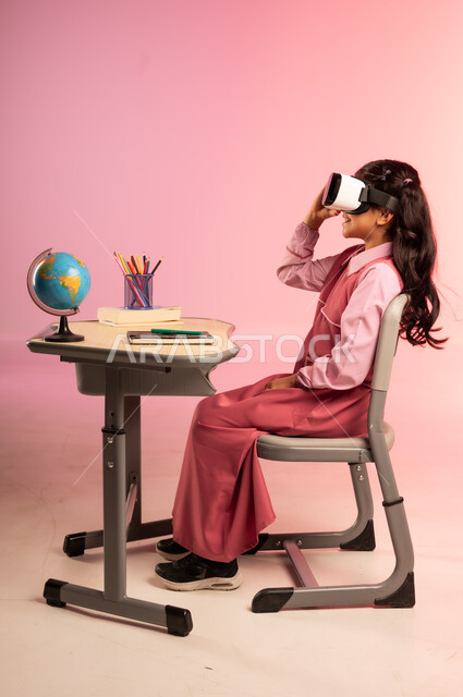  الجد والاجتهاد للوصول إلى النجاح والتفوق، موسم العودة إلى المدارس، بورتريه من الجانب لطالبة عربية خليجية سعودية ترتدي الزي المدرسي وتضع نظارة في آر تجلس في مقعدها وتتفاعل مع الواقع الافتراضي، قرطاسية مدرسية على الطاولة المكتبية، خلفية ملونة