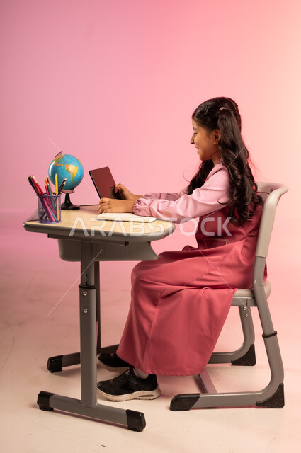 تطور التعليم في مدارس السعودية، الاجتهاد للوصول إلى النجاح والتفوق، موسم العودة إلى المدرسة، بورتريه من الجانب لطالبة عربية خليجية سعودية ترتدي الزي المدرسي وتمسك بالجهاز اللوحي وتتفاعل مع التعليم الالكتروني، قرطاسية مدرسية على الطاولة المكتبية، خلفية ملونة