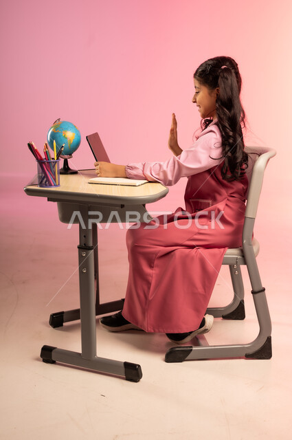 تطور التعليم في مدارس السعودية، الاجتهاد للوصول إلى النجاح والتفوق، موسم العودة إلى المدرسة، بورتريه من الجانب لطالبة عربية خليجية سعودية ترتدي الزي المدرسي وتمسك بالجهاز اللوحي وتتفاعل مع التعليم الالكتروني، قرطاسية مدرسية على الطاولة المكتبية، خلفية ملونة