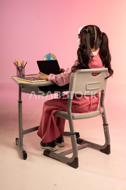 تطور التعليم في مدارس السعودية، الاجتهاد للوصول إلى النجاح والتفوق، موسم العودة إلى المدرسة، بورتريه من الخلف لطالبة عربية خليجية سعودية ترتدي الزي المدرسي وتمسك بالجهاز اللوحي وتتفاعل مع التعليم الالكتروني، قرطاسية مدرسية على الطاولة المكتبية، خلفية ملونة