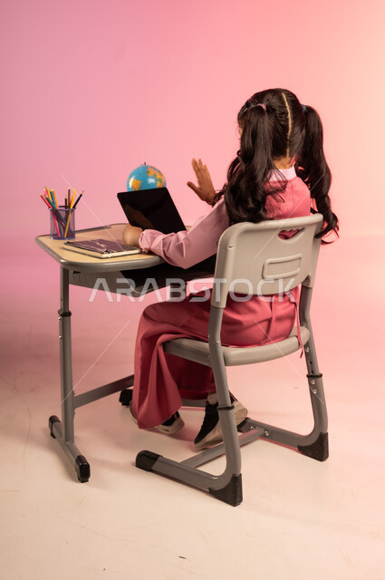 تطور التعليم في مدارس السعودية، الاجتهاد للوصول إلى النجاح والتفوق، موسم العودة إلى المدرسة، بورتريه من الخلف لطالبة عربية خليجية سعودية ترتدي الزي المدرسي وتمسك بالجهاز اللوحي وتتفاعل مع التعليم الالكتروني، قرطاسية مدرسية على الطاولة المكتبية، خلفية ملونة