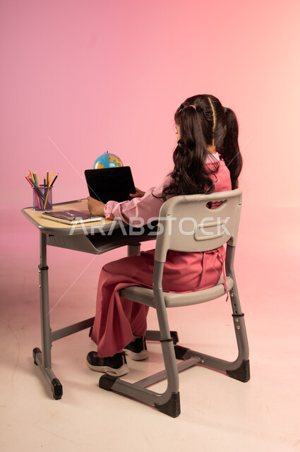 تطور التعليم في مدارس السعودية، الاجتهاد للوصول إلى النجاح والتفوق، موسم العودة إلى المدرسة، بورتريه من الخلف لطالبة عربية خليجية سعودية ترتدي الزي المدرسي وتمسك بالجهاز اللوحي وتتفاعل مع التعليم الالكتروني، قرطاسية مدرسية على الطاولة المكتبية، خلفية ملونة