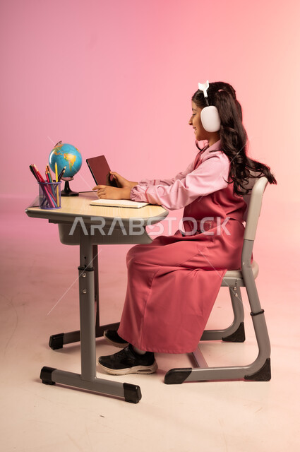 طرائق التعليم المتطورة في مدارس المملكة العربية السعودية، موسم العودة للمدرسة، بورتريه من الجانب لطالبة عربية خليجية سعودية ترتدي الزي المدرسي وسماعات الهيدفون تمسك بالجهاز اللوحي وتتفاعل مع التعليم الالكتروني، قرطاسية مدرسية على الطاولة المكتبية، خلفية ملونة