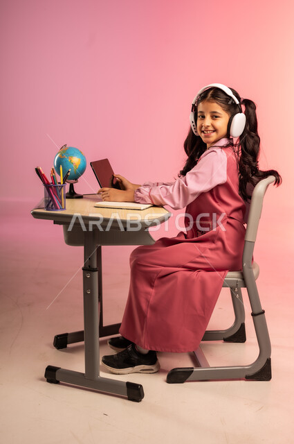 طرائق التعليم المتطورة في مدارس المملكة العربية السعودية، موسم العودة للمدرسة، بورتريه من الجانب لطالبة عربية خليجية سعودية ترتدي الزي المدرسي وسماعات الهيدفون تمسك بالجهاز اللوحي وتتفاعل مع التعليم الالكتروني، قرطاسية مدرسية على الطاولة المكتبية، خلفية ملونة