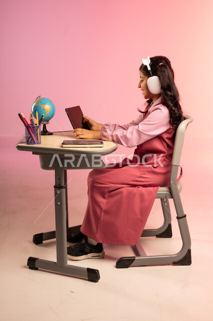 طرائق التعليم المتطورة في مدارس المملكة العربية السعودية، موسم العودة للمدرسة، بورتريه من الجانب لطالبة عربية خليجية سعودية ترتدي الزي المدرسي وسماعات الهيدفون تمسك بالجهاز اللوحي وتتفاعل مع التعليم الالكتروني، قرطاسية مدرسية على الطاولة المكتبية، خلفية ملونة
