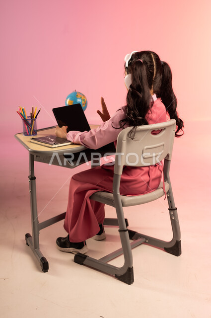 طرائق التعليم المتطورة في مدارس المملكة العربية السعودية، موسم العودة للمدرسة، بورتريه من الخلف لطالبة عربية خليجية سعودية ترتدي الزي المدرسي وسماعات الهيدفون تمسك بالجهاز اللوحي وتتفاعل مع التعليم الالكتروني، قرطاسية مدرسية على الطاولة المكتبية، خلفية ملونة