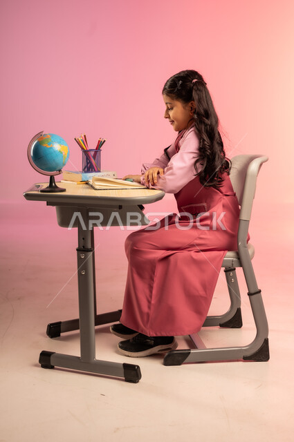 Education and teaching in Saudi Arabia schools, back to school season, side portrait of a Saudi Arabian Gulf Arab female student wearing the official school uniform reviewing lessons and doing homework, school stationery and globe on the office table, colorful background