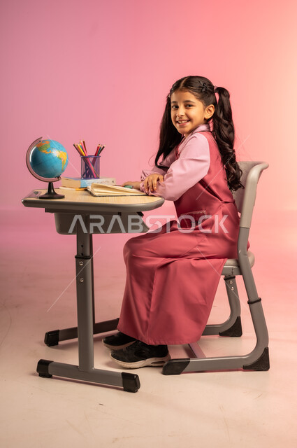 التعليم والتدريس في مدارس المملكة العربية السعودية، موسم العودة للمدرسة، بورتريه من الجانب لطالبة عربية خليجية سعودية ترتدي الزي المدرسي الرسمي وتقوم بمراجعة الدروس وحل الواجبات، قرطاسية مدرسية ومجسم كرة أرضية على الطاولة المكتبية، خلفية ملونة