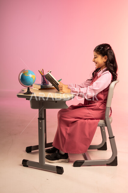 التعليم والتدريس في مدارس المملكة العربية السعودية، موسم العودة للمدرسة، بورتريه من الجانب لطالبة عربية خليجية سعودية ترتدي الزي المدرسي الرسمي وتقوم بمراجعة الدروس وحل الواجبات، قرطاسية مدرسية ومجسم كرة أرضية على الطاولة المكتبية، خلفية ملونة