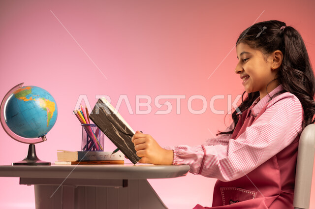 التعليم والتدريس في مدارس المملكة العربية السعودية، موسم العودة للمدرسة، بورتريه من الجانب لطالبة عربية خليجية سعودية ترتدي الزي المدرسي الرسمي وتقوم بمراجعة الدروس وحل الواجبات، قرطاسية مدرسية وجسم كرة أرضية على الطاولة المكتبية، خلفية ملونة
