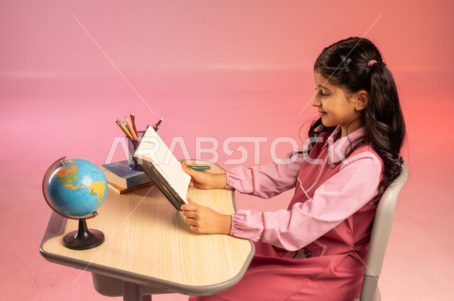 التعليم والتدريس في مدارس المملكة العربية السعودية، موسم العودة للمدرسة، بورتريه من الجانب لطالبة عربية خليجية سعودية ترتدي الزي المدرسي الرسمي وتقوم بمراجعة الدروس وحل الواجبات، قرطاسية مدرسية وجسم كرة أرضية على الطاولة المكتبية، خلفية ملونة