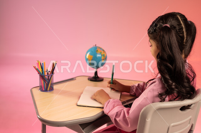  قرطاسية مدرسية ومجسم كرة أرضية على الطاولة المكتبية، التعليم والتدريس في مدارس المملكة العربية السعودية، موسم العودة للمدرسة، بورتريه من الخلف لطالبة عربية خليجية سعودية ترتدي الزي المدرسي الرسمي تجلس في مقعدها وتقوم بمراجعة الدروس وحل الواجبات، خلفية ملونة