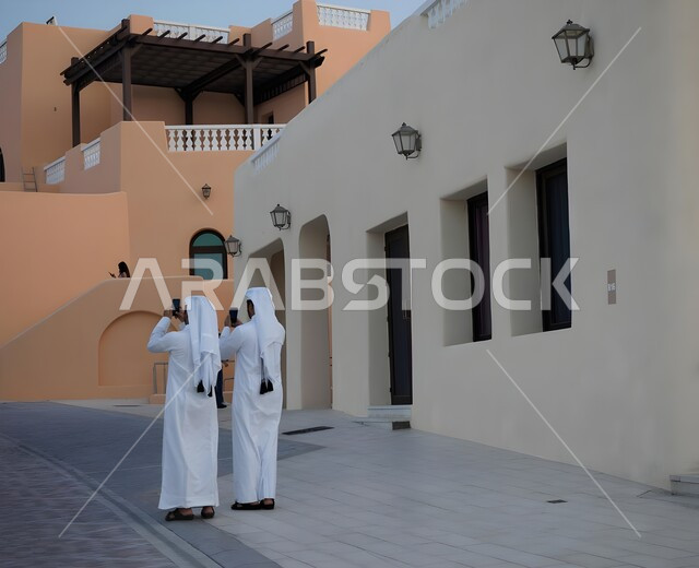 الفن الهندسي المعماري على الطراز الحديث، صورة من الخلف لرجلان عربيان سعوديات يرتديان الثوب التقليدي والشماغ يحملان الهاتف المحمول ويقومان بالتقاط الصور في المدينة، اجراء بث مباشر عبر الانترنت، تصوير اللحظات التذكارية، صورة الجسم بالطول الكامل