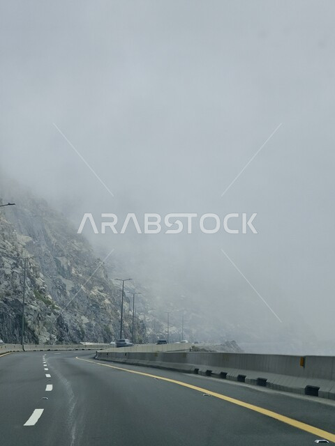 الطبيعة والبيئة الجبلية والمرتفعات في المملكة العربية السعودية، حركة مرور وسير السيارات على طريق الهدا في مدينة الطائف، طريق اسفلتي معبد ملتوي وعر وسط الجبال الصخرية في منطقة جبال الهدا، الجو الضبابي المغطى بالسحب البيضاء في السفوح والمنحدرات وقمم الجبال