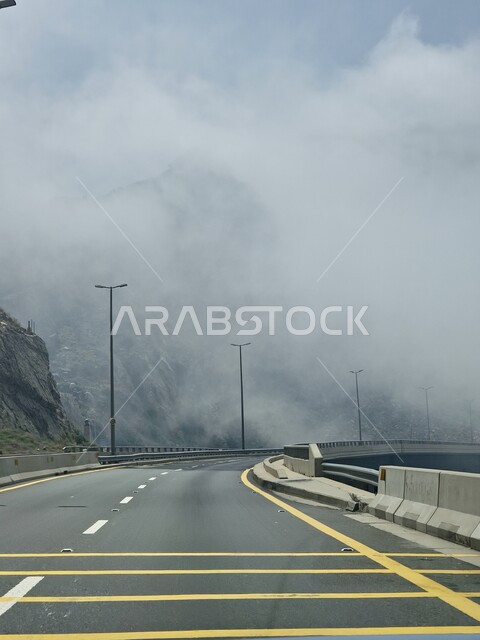 الطبيعة والبيئة الجبلية والمرتفعات في المملكة العربية السعودية، حركة مرور وسير السيارات على طريق الهدا في مدينة الطائف، طريق اسفلتي معبد ملتوي وعر وسط الجبال الصخرية في منطقة جبال الهدا، الجو الضبابي المغطى بالسحب البيضاء في السفوح والمنحدرات وقمم الجبال