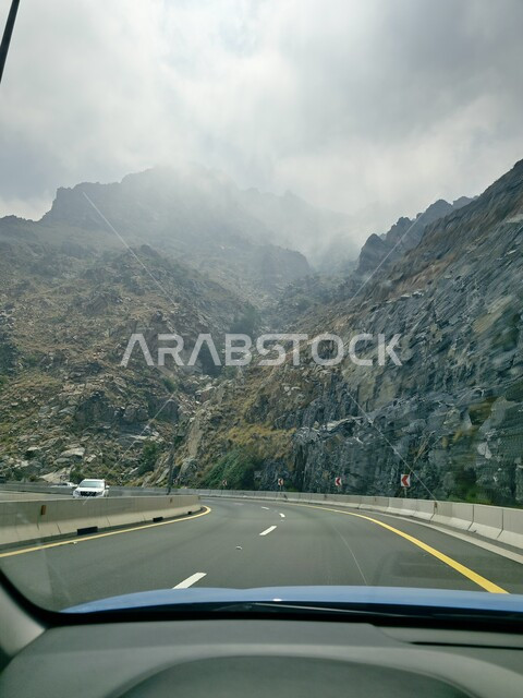 الطبيعة والبيئة الجبلية والمرتفعات في المملكة العربية السعودية، حركة مرور وسير السيارات على طريق الهدا في مدينة الطائف، طريق اسفلتي معبد ملتوي وعر وسط الجبال الصخرية في منطقة جبال الهدا، الجو الضبابي المغطى بالسحب البيضاء في السفوح والمنحدرات وقمم الجبال