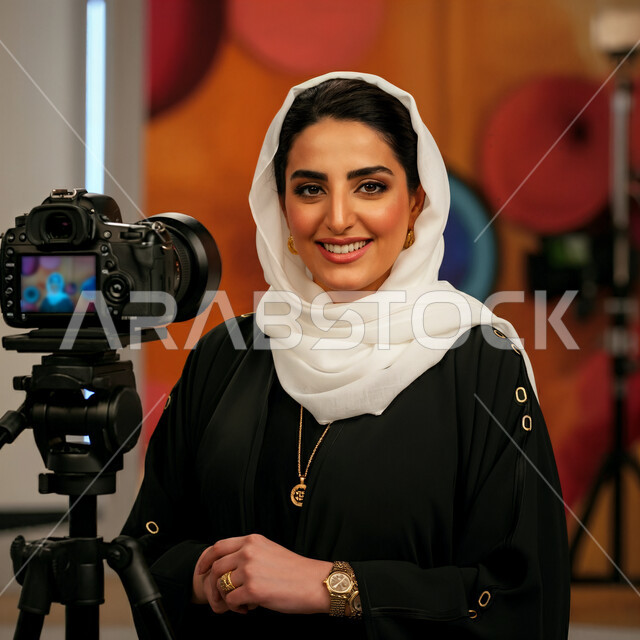 ملابس محجبات عصرية راقية، الاناقة والاهتمام بالمظهر الخارجي، صورة مقربة لشابة عربية خليجية سعودية محجبة مبتسمة تنظر الى الكاميرا بايماءات السرور والسعادة، الوقوف بجوار كاميرا تصوير احتارفية, مفهوم الانوثة والنعومة، تعابير الثقة بالنفس