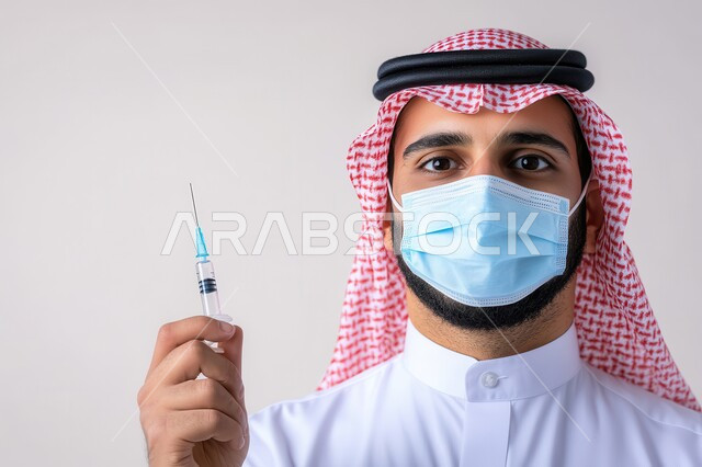 جرعة لتفادي الإصابة بفايروس كورونا, بورتريه مقرب لشاب عربي خليجي سعودي يرتدي الثوب التقليدي و الشماغ يضع القناع الطبي  يمسك بيده جرعة اللقاح، إتباع الإجراءات الاحترازية للوقاية من كوفيد 19, خلفية رمادية