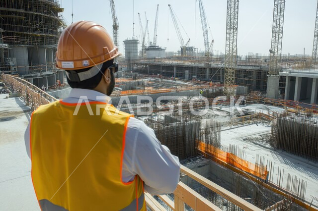 تطور ونمو القطاع الهندسي في المملكة، الاشراف على المشاريع في موقع العمل، صورة من الخلف لمهندس مدني عربي خليجي سعودي يرتدي سترة وخوذة الحماية، مهن ووظائف هندسية سعودية، الات الحفر والبناء في مقر الإنشاءات
