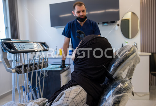العمل في مجال جراحة الفم والفكين، طبيب أسنان عربي خليجي سعودي يرتدي اليونيفورم والقفازات والكمامة الطبية يقوم بتشخيص وشرح الحالة للمريضة المستلقية على كرسي العلاج، معالجة التسوس وتركيب الحشوات، تقديم الخدمات الصحية، أهمية زيارة العيادة السنية بشكل دوري