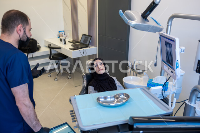 العمل في مجال جراحة الفم والفكين، طبيب أسنان عربي خليجي سعودي يرتدي اليونيفورم والقفازات والكمامة الطبية يقوم بتشخيص وشرح الحالة للمريضة المستلقية على كرسي العلاج، معالجة التسوس وتركيب الحشوات، تقديم الخدمات الصحية، أهمية زيارة العيادة السنية بشكل دوري