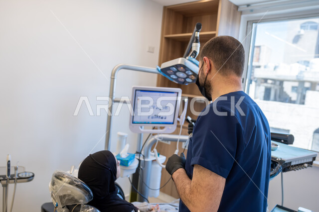 دمج التقنيات الحديثة والتكنولوجيا في مجال الطب، العمل في قطاع جراحة الفم والفكين، طبيب أسنان عربي خليجي سعودي يرتدي اليونيفورم والقفازات والكمامة الطبية يقوم بشرح الحالة الظاهرة على شاشة العرض للمريضة المستلقية على كرسي العلاج، معالجة التسوس وتركيب الحشوات
