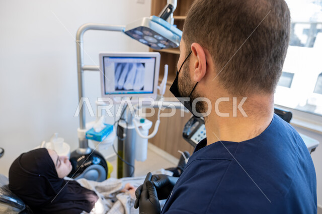 دمج التقنيات الحديثة والتكنولوجيا في مجال الطب، العمل في قطاع جراحة الفم والفكين، طبيب أسنان عربي خليجي سعودي يرتدي اليونيفورم والقفازات والكمامة الطبية يقوم بشرح الحالة الظاهرة على شاشة العرض للمريضة المستلقية على كرسي العلاج، معالجة التسوس وتركيب الحشوات