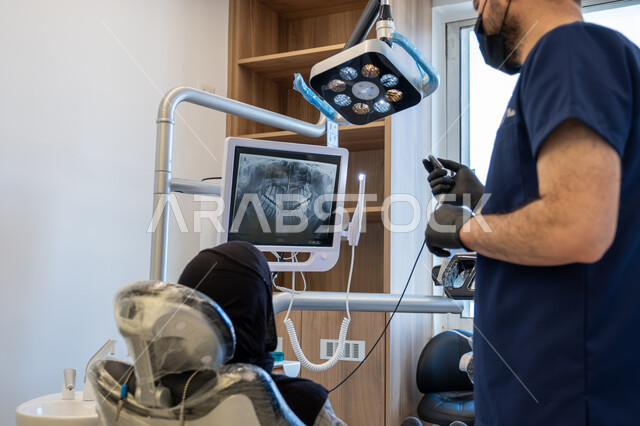 دمج التقنيات الحديثة والتكنولوجيا في مجال الطب، العمل في قطاع جراحة الفم والفكين، طبيب أسنان عربي خليجي سعودي يرتدي اليونيفورم والقفازات والكمامة الطبية يقوم بشرح الحالة الظاهرة على شاشة العرض للمريضة المستلقية على كرسي العلاج، معالجة التسوس وتركيب الحشوات