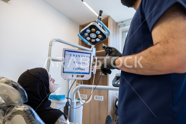 دمج التقنيات الحديثة والتكنولوجيا في مجال الطب، العمل في قطاع جراحة الفم والفكين، طبيب أسنان عربي خليجي سعودي يرتدي اليونيفورم والقفازات والكمامة الطبية يقوم بشرح الحالة الظاهرة على شاشة العرض للمريضة المستلقية على كرسي العلاج، معالجة التسوس وتركيب الحشوات