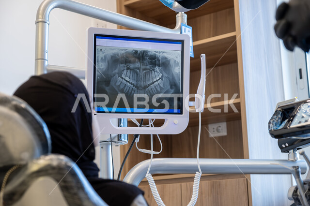 دمج التقنيات الحديثة والتكنولوجيا في مجال الطب، العمل في قطاع جراحة الفم والفكين، طبيب أسنان عربي خليجي سعودي يرتدي اليونيفورم والقفازات والكمامة الطبية يقوم بشرح الحالة الظاهرة على شاشة العرض للمريضة المستلقية على كرسي العلاج، معالجة التسوس وتركيب الحشوات
