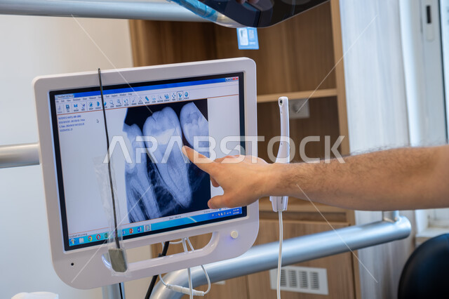 الاستفادة من التصوير الشعاعي والتقنيات الحديثة والتكنولوجيا في مجال الطب، العمل في قطاع جراحة الفم واللثة، طبيب أسنان عربي خليجي سعودي يقوم بالإشارة إلى الحالة المرضية الظاهرة على شاشة العرض لصورة الأشعة للفكين، الحاجة لمعالجة التسوس والالتهاب وتركيب الحشوات