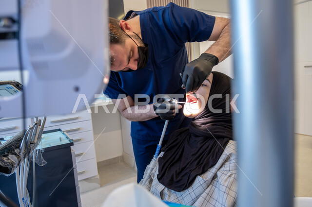العمل في قطاع جراحة الفم والفكين، صورة مقربة لطبيب أسنان عربي خليجي سعودي يرتدي اليونيفورم والقفازات والكمامة الطبية يقوم بمعالجة النخور والتسوس للمريضة المستلقية على كرسي العلاج، استخدام الأدوات المعقمة والتقنيات والأجهزة الحديثة