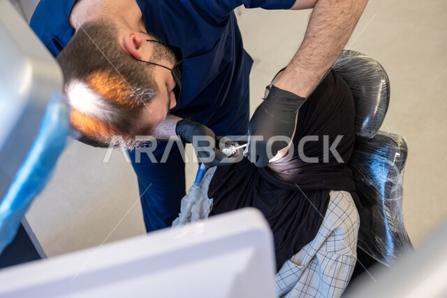 العمل في قطاع جراحة الفم والفكين، صورة مقربة لطبيب أسنان عربي خليجي سعودي يرتدي اليونيفورم والقفازات والكمامة الطبية يقوم بمعالجة النخور والتسوس للمريضة المستلقية على كرسي العلاج، استخدام الأدوات المعقمة والتقنيات والأجهزة الحديثة
