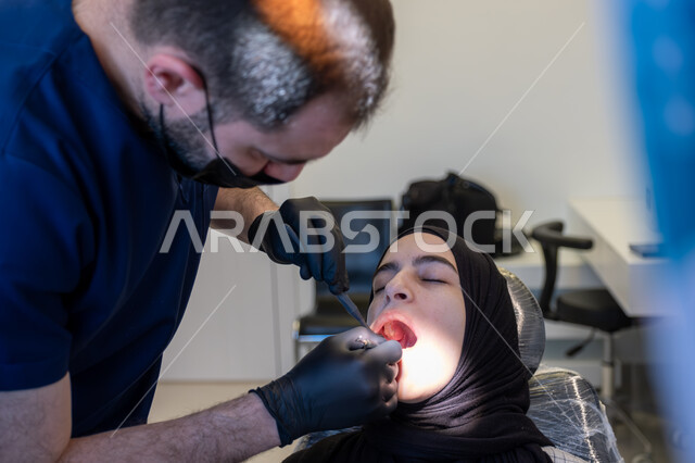العمل في قطاع جراحة الفم والفكين، صورة مقربة لطبيب أسنان عربي خليجي سعودي يرتدي اليونيفورم والقفازات والكمامة الطبية يقوم بمعالجة النخور والتسوس للمريضة المستلقية على كرسي العلاج، استخدام الأدوات المعقمة والتقنيات والأجهزة الحديثة