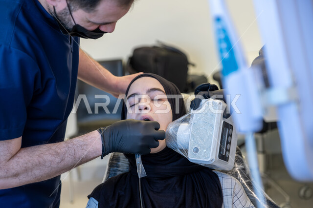 Using sterile tools, modern techniques and devices, working in the field of oral and maxillofacial surgery, close-up image of a Saudi Arabian Gulf Arab dentist wearing a uniform, medical gloves and a mask treating cavities and decay for a patient lying on a treatment chair in a dental clinic