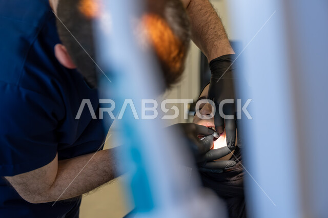 Using sterile tools, modern techniques and devices, working in the field of oral and maxillofacial surgery, close-up image of a Saudi Arabian Gulf Arab dentist wearing a uniform, medical gloves and a mask treating cavities and decay for a patient lying on a treatment chair in a dental clinic