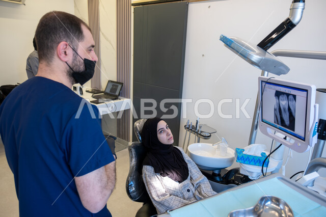 تقديم الاستشارات والنصائح الطبية، العمل في مجال جراحة الفم والفكين، طبيب أسنان عربي خليجي سعودي يرتدي اليونيفورم والقفازات والكمامة يقوم بشرح الحالة للمريضة المستلقية على كرسي العلاج في العيادة السنية، استخدام الأدوات المعقمة والتقنيات والأجهزة الحديثة