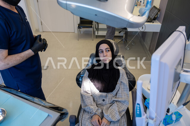 تقديم الاستشارات والنصائح الطبية، العمل في مجال جراحة الفم والفكين، صورة من الأعلى لمريضة مستلقية على كرسي العلاج في العيادة السنية وطبيب أسنان عربي خليجي سعودي يرتدي اليونيفورم والقفازات يقوم بشرح الحالة لها، استخدام الأدوات المعقمة والتقنيات والأجهزة الحديثة